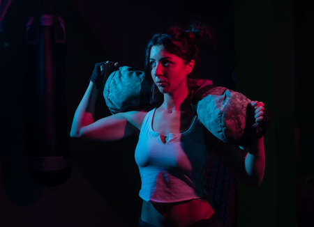 Young sport woman holding a heavy fitness bag for workout in neon gradient blue red light over a dark background. Functional Training Conceptの写真素材
