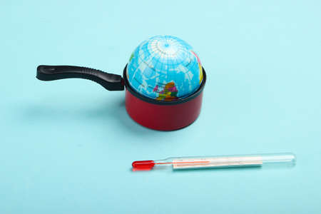 Global warming concept. Mini globe in a toy pan and thermometer on blue background. Minimalism. Climatic problems of our time.の写真素材