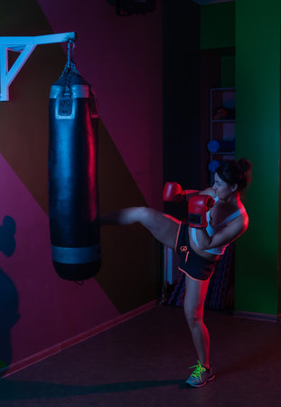 Female boxer in boxing gloves kicking a punching bag in red blue neon light over dark backgroundの写真素材