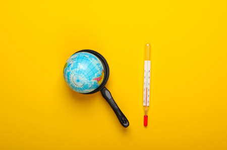 Global warming concept. Mini globe in a toy pan and thermometer on yellow background. Minimalism....