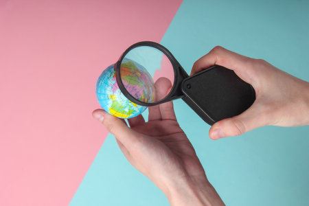 Female hand holds a globe with a magnifying glass on blue-pink pastel background. Travel, Tourism...