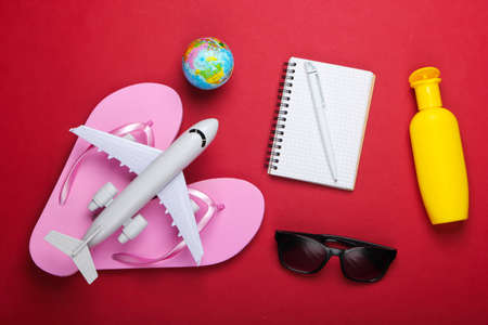 Planning Travel and beach vacation. Summer background. Flip flops, airplane figurine, globe, sunglasses and sunblock, notebook on red background. Top view. Flat layの写真素材