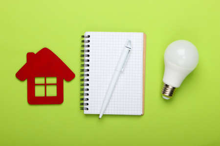 Planning energy saving. House figurine and energy-saving light bulb, notebook on green background. Top viewの写真素材