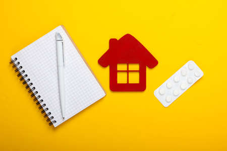 Medicine at home. Home treatment. House figurine, blister of pills, notebook on yellow background. Top viewの写真素材