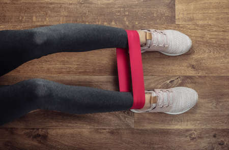 Female legs in sports leggings and sneakers doing exercises with fitness elastic on a wooden floor....