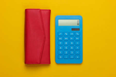 Manage family budget. Shopping costs. Blue calculator with red leather wallet on yellow background. Top viewの写真素材