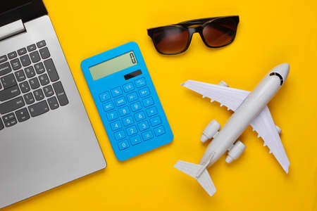 Calculation of the cost of tourism or emigration. Online booking. Laptop, plane, calculator and sunglasses on a yellow background. Top viewの写真素材