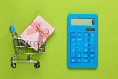 Calculator and shopping trolley with gift box on green background. Calculation of the value of the...