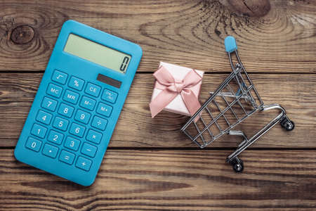 Shopping trolley with gift box and calculator on a wooden table. Holiday shopping cost calculationの写真素材