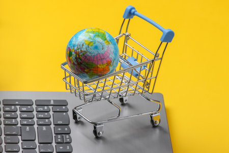 Global online supermarket. Shopping trolley with globe on laptop keyboard. Yellow background