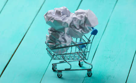 Shopping trolley with crumpled paper balls on a blue wooden background.の写真素材