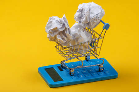 Calculator and shopping trolley with a ball of crumpled paper on yellow background. Top viewの写真素材