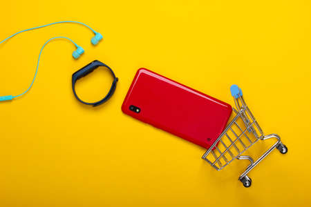 Modern gadgets. Shopping concept. Smartphone and smart bracelet, vacuum earphones and mini supermarket trolley on yellow background. Top viewの写真素材