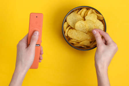 Movie time. Hand holding Tv remotes and bowl with chips on yellow background. Top viewの写真素材