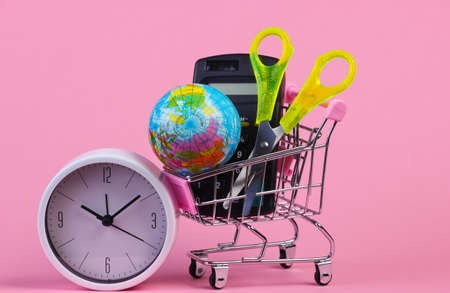 School supplies in a shopping trolley. Calculator, globe, scissors in supermarket cart, clock on...