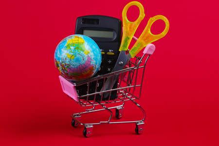 School supplies in a shopping trolley. Calculator, globe, scissors in supermarket cart on red background. back to schoolの写真素材