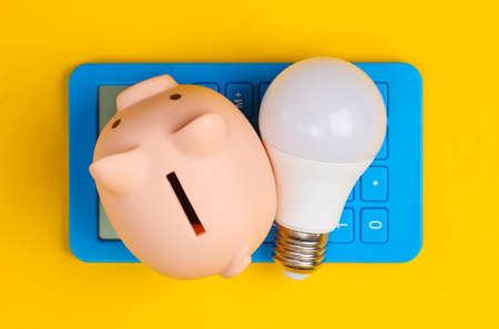 Piggy bank and led light bulb, calculator on yellow background. Energy saving. Minimalistic studio...