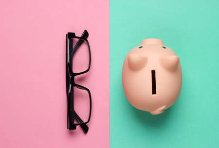 Piggy bank and glasses on pink blue background. Minimalistic studio shot. Overhead view. Flat lay.の写真素材