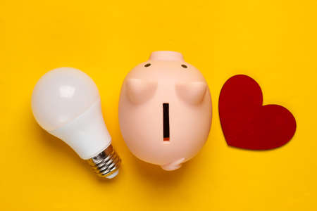 Piggy bank and led light bulb, heart on yellow background. Energy saving. Minimalistic studio shot. Overhead view. Flat lay.の写真素材