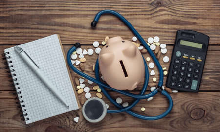 Medicine concept. Medical budget. Stethoscope with piggy bank, pills, calculator, notebook on wooden background. Top view. Flat layの写真素材
