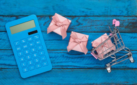Shopping concept, economy. Supermarket trolley with gift boxes and calculator on blue wooden...