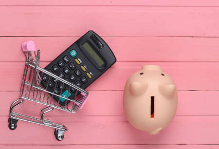 Shopping concept, economy. Piggy bank with supermarket trolley, calculator on pink wooden...