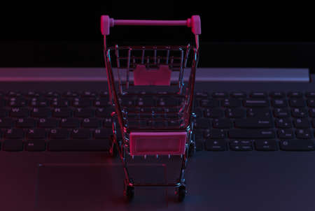 Mini supermarket trolley on laptop keyboard. Neon gradient red-blue, ultraviolet light. Online shoppingの写真素材