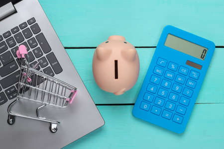 Online shopping ideas. Laptop with piggy bank, supermarket trolley, calculator on a blue wooden background. Saving concept. Top viewの写真素材