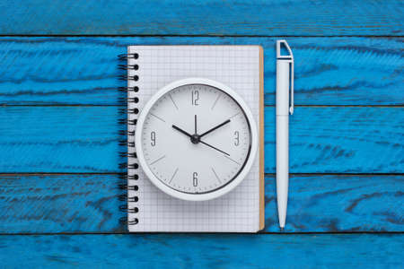 White clock and notebook on blue wooden background. Minimalistic studio shot. Top viewの写真素材
