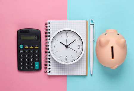 Time to invest. White clock, notebook, piggy bank and calculator on pink blue pastel background. Minimalistic studio shot. Top viewの写真素材