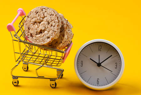 Diet concept. Shopping trolley with whole grain crispbreads and clock on yellow background