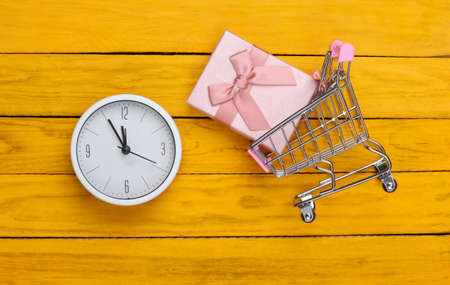 Christmas, New Year shopping. Mini supermarket trolley with gift box and clock on yellow wooden...