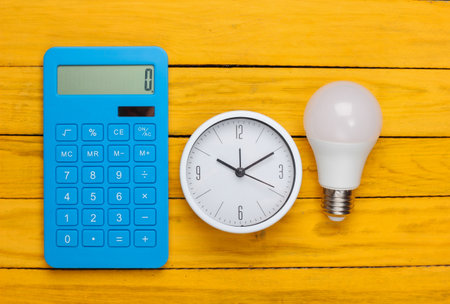 Clock with light bulb, calculator on a yellow wooden background. Minimalism. Top viewの写真素材