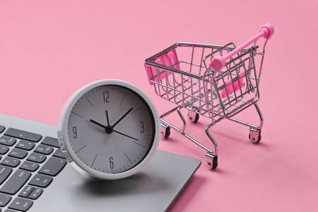 Online shopping time. Computer keyboard with clock and supermarket trolley on pink background.の写真素材