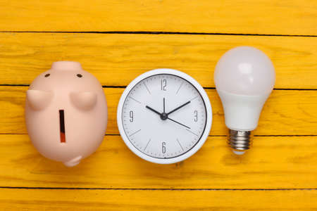Clock with light bulb, piggy bank on a yellow wooden background. Minimalism. Top viewの写真素材