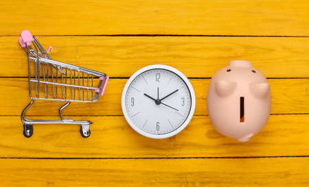 Shopping time. Supermarket trolley with piggy bank, clock on a yellow wooden background. Minimalism. Top viewの写真素材