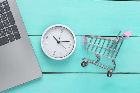 Online shopping time. Computer keyboard with clock and supermarket trolley on blue wooden...