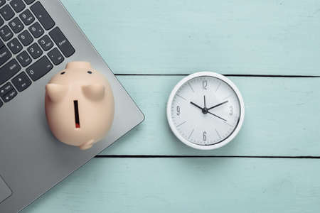 Time to invest, online business. Piggy bank with clock, laptop keyboard on blue wooden background....