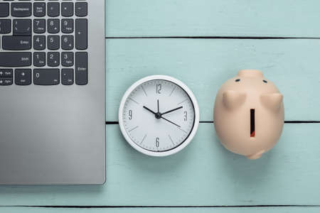 Time to invest, online business. Piggy bank with clock, laptop keyboard on blue wooden background....