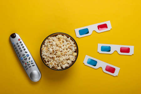 Movie time. Stereoscopic anaglyph disposable paper 3d glasses, tv remote and popcorn bowl on yellow background. Top viewの写真素材