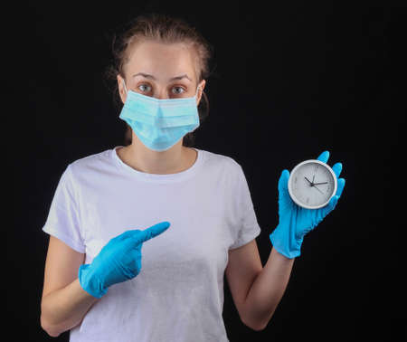 Woman in a medical protective mask, gloves hold white clock on a black background.の写真素材