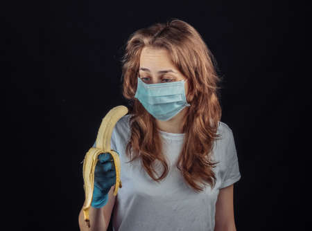 Woman in protective medical mask and gloves tries to eat a banana on black backgroundの写真素材