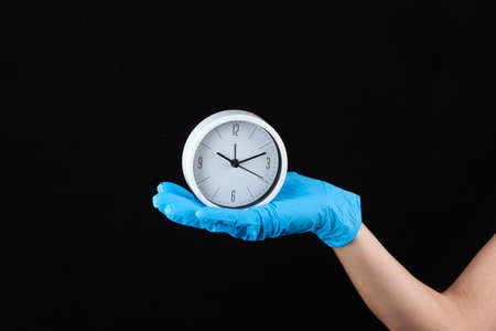 Hand in blue medical gloves hold white clock on a black background.の写真素材