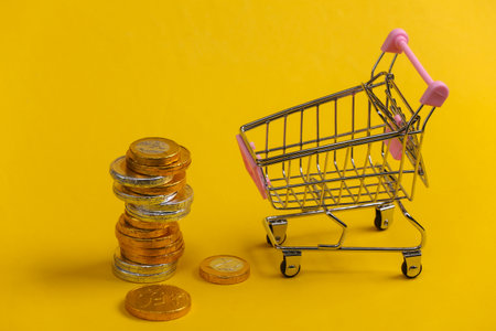 Shopping theme. Mini Supermarket trolley with coins on yellow background.