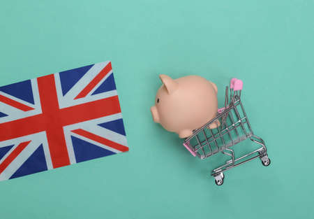 Shopping trolley with piggy bank and British flag on mint green background.