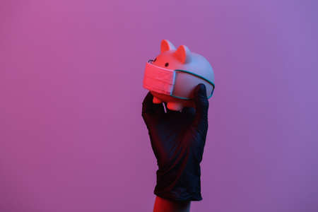 A hand in a glove holds a piggy bank with a medical mask. Red-blue neon gradient lightの写真素材