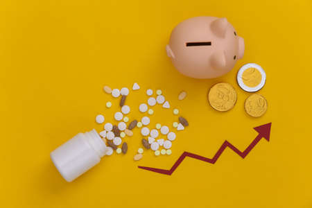 Bottle of pills with arrow tending up, piggy bank with coins on yellow background. Top viewの写真素材