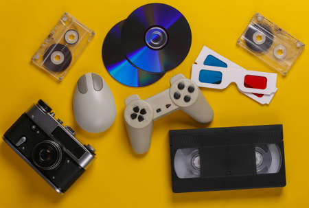 Retro entertainment. Attributes, gadgets 80s. Keyboard, pc mouse, compact discs, gamepad, anaglyph glasses, audio and video cassette, camera on yellow background. Top view. Flat layの写真素材