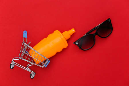 Shopping trolley with sunblock bottle, sunglasses on red background. skin protection. beach...