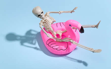 Toy skeleton with inflatable flamingo on blue bright background. Halloween theme. Beach vacation...